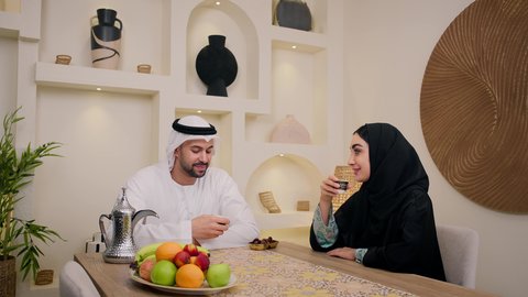 Combining customs and traditions with the warmth of everyday life, an atmosphere of authentic Arab hospitality that reflects the Gulf heritage, an Arab Gulf Emirati couple sitting together in a warm home atmosphere, an Emirati man wearing a kandura and ghutra, pouring Arabic coffee into a cup for his wife, friendly moments full of respect and love