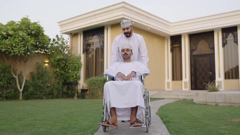 الاهتمام بالجانب النفسي والجسدي للمقعدين,  نشاط مسلي في إحدى الشاليهات الراقية في سلطنة عمان, تبادل الحوار بإيماءات الاندماج، شاب من ذوي الهمم عربي خليجي عماني يرتدي الثوب التقليدي  يجلس على كرسي متحرك يقضي وقت ممتع مع صديقه في الهواء الطلق