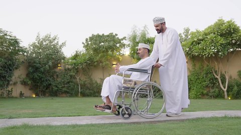 نشاط مسلي في إحدى الشاليهات الراقية في سلطنة عمان, تبادل الحوار بإيماءات الاندماج، الاهتمام بالجانب النفسي والجسدي للمقعدين, شاب من ذوي الهمم عربي خليجي عماني يرتدي الثوب التقليدي  يجلس على كرسي متحرك يقضي وقت ممتع مع صديقه في الهواء الطلق