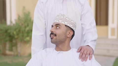 تبادل الحوار بإيماءات الاندماج، نشاط مسلي في إحدى الشاليهات الراقية في سلطنة عمان, الاهتمام بالجانب النفسي والجسدي للمقعدين, شاب من ذوي الهمم عربي خليجي عماني يرتدي الثوب التقليدي  يجلس على كرسي متحرك يقضي وقت ممتع مع صديقه في الهواء الطلق