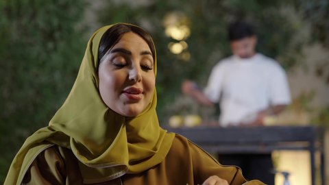 Chatting and exchanging conversations between friends with gestures of integration, a close-up of an Omani Gulf Arab woman wearing an abaya eating fruit before dinner, entertaining events and activities, outings and entertainment in a luxury tourist chalet during the holiday, barbecue background