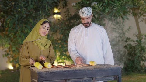 Cooperation and participation in preparing food, chatting and exchanging conversations between spouses with gestures of integration, a close-up of an Omani Gulf Arab woman wearing an abaya participating with her husband in the process of grilling food, entertaining events and activities, outings and entertainment in a high-end tourist chalet during the holiday, barbecue background