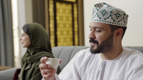التنزه والتسلية والترفيه في شاليه سياحية في عمان، اجتماع الأهل والأصدقاء في الهواء الطلق، تصوير مقرب لرجل عربي خليجي عماني يرتدي الثوب التقليدي يمسك فنجان القهوة يقوم بالتحدث مع أصدقائه بإيماءات الاندماج، قضاء وقت ممتع ومسلي مع مجموعة من الأصدقاء