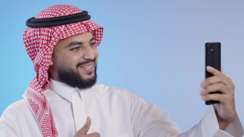 إجراء مكالمة فيديو بإيماءات السرور عبر الانترنت, رجل عربي خليجي سعودي مبتسم يرتدي الثوب التقليدي والشماغ يستخدم الهاتف المحمول, التواصل مع الأصدقاء والأقارب عبر التطبيقات والتقنيات الحديثة والمتطورة