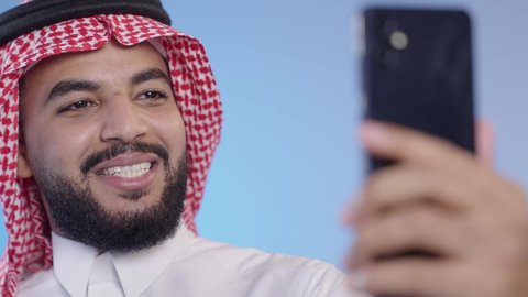 إجراء مكالمة فيديو بايماءات البشاشة، تعابير الفرح والسرور، رجل عربي خليجي سعودي مبتسم يرتدي الثوب التقليدي و الشماغ يمسك الهاتف المحمول, استخدام الأجهزة التقنية الحديثة