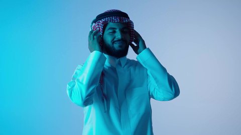 ايماءات وجه ويدين تدل على السعاده والتفاعل مع الأغاني, الاستمتاع بتقنية الهيدفون، رجل عربي خليجي سعودي مبتسم يرتدي الثوب التقليدي و الشماغ  و يستخدم الجوال والأجهزة التكنولوجية