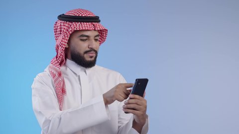 استخدام الهاتف المحمول لإنجاز المهام، تصفح وسائل التواصل الاجتماعي, استخدام الأجهزة التقنية الحديثة و المتطورة, رجل عربي خليجي سعودي مبتسم يرتدي الثوب التقليدي و الشماغ يمسك الجوال بيده