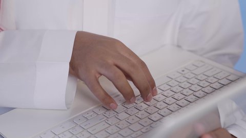 النقر على لوحة المفاتيح, إدارة الأعمال عن بعد, رجل عربي خليجي سعودي يرتدي الثوب التقليدي يقوم بانجاز المهام عبر اللابتوب, تصفح الانترنت وبرامج التواصل الاجتماعي