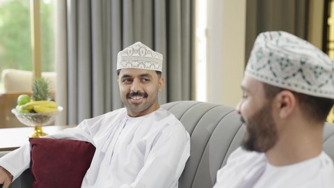 تعارف وصداقة وبناء علاقات اجتماعية، تبادل الاخبار والاحاديث، جلسة وناسة يوم الإجازة، رجلين عربيين خليجيين عمانيين يرتديان الدشداشه والكمة يجلسان في شاليه راقي بإيماءات السعادة والسرور