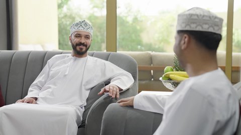 تعارف وصداقة وبناء علاقات اجتماعية، تبادل الاخبار والاحاديث، جلسة وناسة يوم الإجازة، رجلين عربيين خليجيين عمانيين يرتديان الدشداشه والكمة يجلسان في شاليه راقي بإيماءات السعادة والسرور