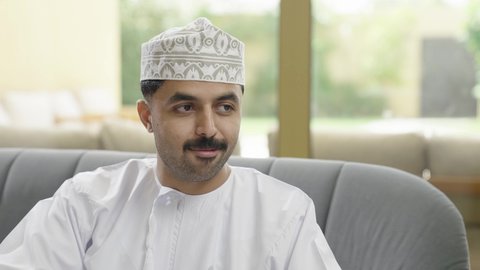 الانصات وسماع سوالف سعيده، تصوير مقرب لرجل عربي خليجي عماني يرتدي الدشداشة والكمة  يجلس على اريكه يقوم بالتحدث مع شخص ما، ايماءات السعادة والسرور، تبادل الاخبار والاحاديث