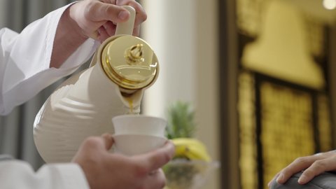 مفهوم حسن الضيافة والكرم، تصوير مقرب ليد رجل عربي خليجي عماني يرتدي الدشداشة يقوم بصب القهوة العربية الساخنة في الفنجان، تبادل الاخبار والاحاديث