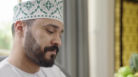 ايماءات السعادة والسرور، الانصات وسماع سوالف سعيده، تصوير مقرب لرجل عربي خليجي عماني يرتدي الدشداشة والكمة  يجلس على اريكه يقوم بالتحدث مع شخص ما، تبادل الاخبار والاحاديث