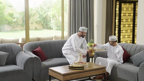 جلسة وناسة في عطل نهاية الاسبوع، تقديم القهوة والحلويات العربية، حسن الاستقبال وكرم الضيافة، جلسة هادئة في احدى شاليهات عمان، صديقان عربيان خليجيان عمانيان يرتديان الدشداشة والكمة يجلسان على الاريكة يقومان بتبادل الأخبار والأحاديث