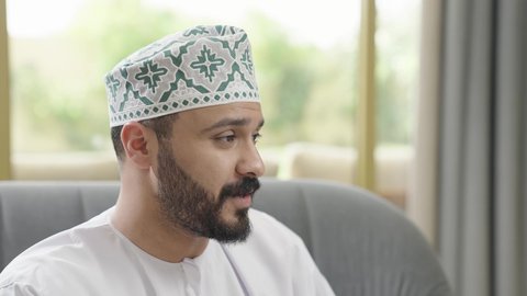 ايماءات السعادة والسرور، الانصات وسماع سوالف سعيده، تصوير مقرب لرجل عربي خليجي عماني يرتدي الدشداشة والكمة  يجلس على اريكه يقوم بالتحدث مع شخص ما، تبادل الاخبار والاحاديث