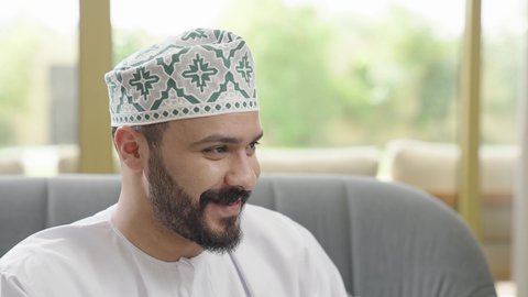 Gestures of happiness and joy, listening and hearing pleasant conversations, a close-up shot of an Arab Gulf Omani man wearing a dishdasha and kumma sitting on a couch talking to someone, exchanging news and discussions.