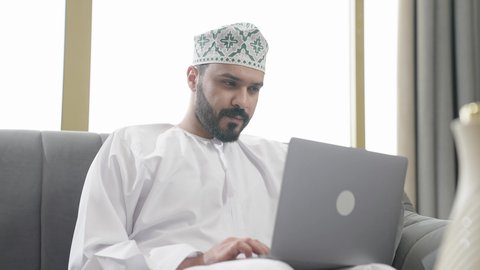 ايماءات الاندماج والتركيز، جهاز تقني حديث ومتطور، إنجاز مهام العمل عن بعد عبر اللابتوب، شاب عربي خليجي عماني مثقف يرتدي الدشداشة والكمة يجلس في إحدى الشاليهات يقوم باستخدام الحاسوب المحمول، تصفح الانترنت ومواقع السوشال ميديا