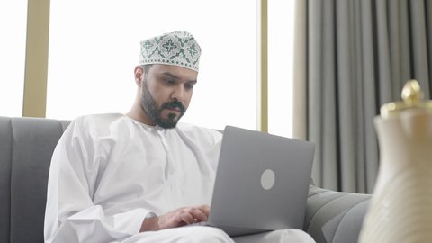 ايماءات الاندماج والتركيز، جهاز تقني حديث ومتطور، إنجاز مهام العمل عن بعد عبر اللابتوب، شاب عربي خليجي عماني مثقف يرتدي الدشداشة والكمة يجلس في إحدى الشاليهات يقوم باستخدام الحاسوب المحمول، تصفح الانترنت ومواقع السوشال ميديا