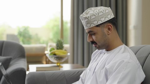 Expressions of happiness and pleasure, a modern and advanced technical device, completing work tasks remotely via laptop, a cultured young Arab Gulf Omani man wearing a dishdasha and a turban sitting in a chalet using a laptop, browsing the Internet and social media sites