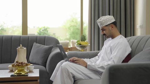 ايماءات الاندماج والتركيز، جهاز تقني حديث ومتطور، إنجاز مهام العمل عن بعد عبر اللابتوب، شاب عربي خليجي عماني مثقف يرتدي الدشداشة والكمة يجلس في إحدى الشاليهات يقوم باستخدام الحاسوب المحمول، تصفح الانترنت ومواقع السوشال ميديا