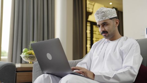 تعابير السعادة والسرور، جهاز تقني حديث ومتطور، إنجاز مهام العمل عن بعد عبر اللابتوب، شاب عربي خليجي عماني مثقف يرتدي الدشداشة والكمة يجلس في إحدى الشاليهات يقوم باستخدام الحاسوب المحمول، تصفح الانترنت ومواقع السوشال ميديا