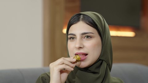 Features of femininity and softness, a smiling Omani Gulf Arab woman wearing a hijab with a colorful abaya sitting on a gray sofa looking at something, a close-up of a smiling Saudi woman eating a date, a local national product