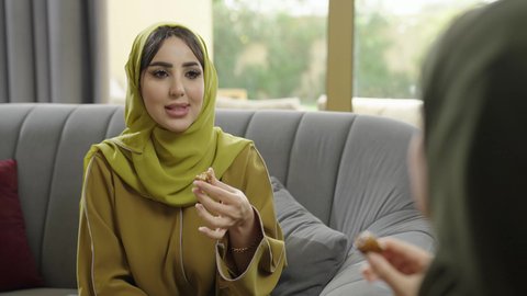التلذذ بالحلويات الشرقية، حسن الضيافة والكرم، بناء علاقات اجتماعية ناجحة، سيدتان عمانيتان تقضيان اوقات ممتعة في إحدى الشاليهات، تصوير مقرب لامرأة عربية خليجية سعودية ترتدي الحجاب والعباءة تجلس على الأريكة مع صديقتها بإيماءات السعادة، تناول القهوة والحلى