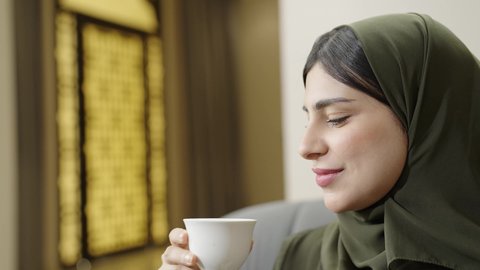 الاستمتاع بتناول فنجان قهوه ساخن، إمرأه عربية خليجية عمانيه ترتدي الحجاب مع عباءة ملونه تجلس علي اريكه رماديه تتحدث مع شخص ما، تصوير مقرب لسيده سعوديه مبتسمه تنظرالي صديقتها تقوم بالاستماع الجيد، مظهرالنعومه والانوثه