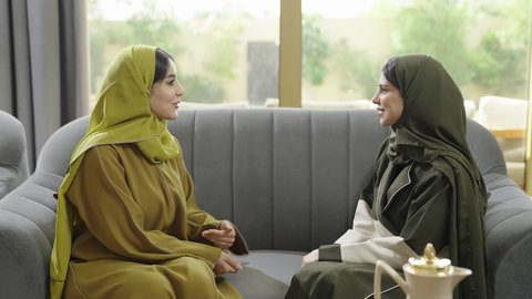 Building successful social relationships, discussions and dialogues in a calm atmosphere, two Omani women spending a pleasant time in a chalet, a Saudi Arabian Gulf woman wearing a hijab and an abaya sitting on the couch with her friend with gestures of happiness, hospitality and generosity, drinking coffee and sweets