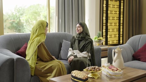 بناء علاقات اجتماعية ناجحة، النقاشات والحوارات في الأجواء الهادئة، سيدتان عمانيتان تقضيان اوقات ممتعة في إحدى الشاليهات، امرأة عربية خليجية سعودية ترتدي الحجاب والعباءة تجلس على الأريكة مع صديقتها بإيماءات السعادة، حسن الضيافة والكرم، تناول القهوة والحلويات