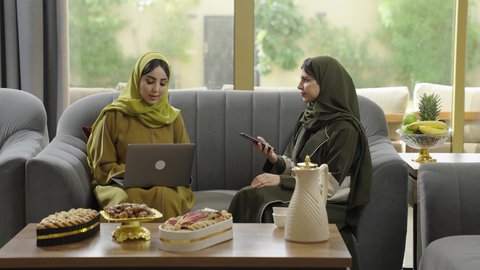استخدام الاجهزه الحديثه، إمرأتان عربيتان خليجيتان عمانيتان مبتسمتان ترتديان الحجاب مع عباءة ملونه تجلسان علي اريكه رماديه تستخدمان احدث التقنيات، سيدتان سعوديتان مبتسمتان تقومان بإنجاز العمل عن بعد، التطور التقني