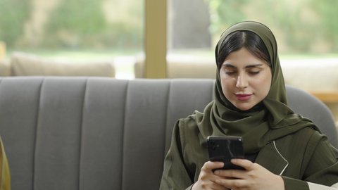 An Omani Gulf Arab woman wearing a hijab with a colorful abaya sitting on a gray sofa, browsing social media on her mobile phone, communicating remotely and clicking on her mobile phone screen, a Saudi woman using a mobile phone