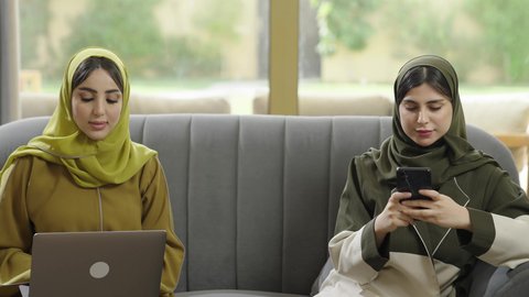استخدام الاجهزه الحديثه، إمرأتان عربيتان خليجيتان عمانيتان مبتسمتان ترتديان الحجاب مع عباءة ملونه تجلسان علي اريكه رماديه تستخدمان احدث التقنيات، سيدتان سعوديتان مبتسمتان تقومان بإنجاز العمل عن بعد، منتج محلي وطني