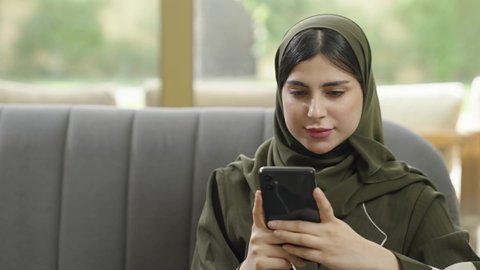 تصفح وسائل التواصل الاجتماعي عبر الجوال, التواصل عن بعد والنقر على شاشة الهاتف المحمول, سيدة عربية خليجية عمانيه ترتدي الحجاب مع عباءة ملونه تجلس علي اريكه رماديه, سيده سعوديه تقوم باستخدام الهاتف المحمول