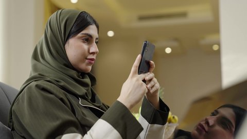 التواصل عن بعد والنقر على شاشة الهاتف المحمول, تصفح وسائل التواصل الاجتماعي عبر الجوال, سيدة عربية خليجية عمانيه ترتدي الحجاب مع عباءة ملونه تجلس علي اريكه رماديه, سيده سعوديه تقوم باستخدام الهاتف المحمول