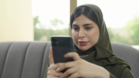 Using a modern technical device, tapping on a mobile phone screen, browsing social media via mobile, an Arab Gulf Omani woman wearing a hijab with a colorful abaya sitting on a gray sofa, a Saudi woman using a mobile phone