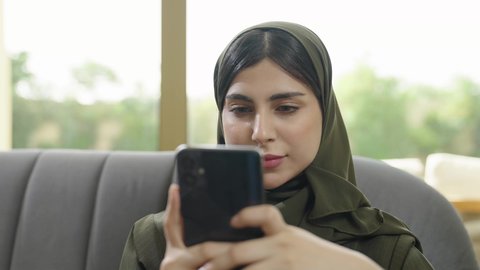 استخدام جهاز تقني حديث، النقر على شاشة الهاتف المحمول, تصفح وسائل التواصل الاجتماعي عبر الجوال, سيدة عربية خليجية عمانيه ترتدي الحجاب مع عباءة ملونه تجلس علي اريكه رماديه, سيده سعوديه تقوم باستخدام الهاتف المحمول