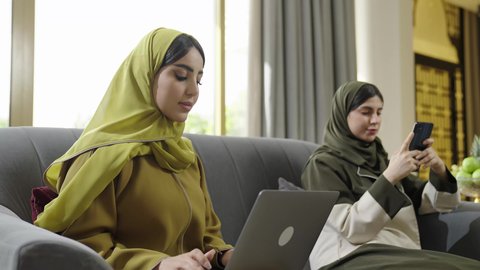 العمل عن بعد، استخدام الاجهزه الحديثه، إمرأتان عربيتان خليجيتان عمانيتان مبتسمتان ترتديان الحجاب مع عباءة ملونه تجلسان علي اريكه رماديه تستخدمان احدث التقنيات، سيدة سعودية تستخدم الحاسب المحمول في انجاز المهام اليومية، الاهتمام بالاناقة والمظهر الخارجي.