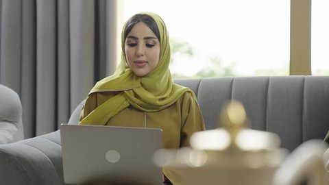 دمج التقنية والتكنولوجيا في الحياة العملية،  سيدة عربية خليجية عمانيه مبتسمة ترتدي العباءة تقوم باستخدام جهاز الكمبيوتربإيماءات البشاشة والسرور, مهن ووظائف نسائيه، إمرأه سعوديه تقوم إنجاز المهام المكتبية من المنزل