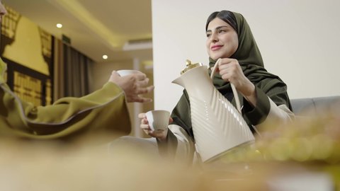 سيده عربيه خليجيه عمانيه ترتدي العباءه والحجاب تقوم بتقديم القهوه العربيه الساخنه, إمرأه سعوديه انيقه تجلس برفقه صديقتها في غرفه المعيشه, جلسه وناسه وتعارف, منتج وطني عربي