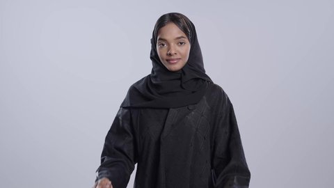 Standing upright and confident, the concept of femininity and softness, interest in one's appearance, portrait of a dark-skinned Saudi Arabian university student wearing an abaya and hijab looking at the camera with gestures of happiness and pleasure, white background