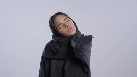 The need to see a doctor and receive treatment. Neck, bone, and spine pain. Facial expressions indicating distress and pain. Portrait of a black, Arab, Saudi university student wearing an abaya and hijab holding her neck with gestures of pain. White background.