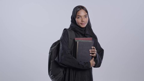 مراجعة الدروس اليومية،التعليم الجامعي في المملكه العربيه السعوديه ،طالبة جامعية سمراء عربيه سعوديه ترتدي العباءه والحجاب تضع الحقيبة على ظهرها ، تحمل بيدها كتب جامعيه،   خلفية بيضاء .