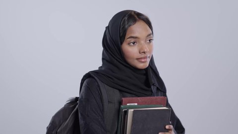 التعليم الجامعي في المملكه العربيه السعوديه ،طالبة جامعية سمراء عربيه سعوديه ترتدي العباءه والحجاب تضع الحقيبة على ظهرها ، تحمل بيدها كتب جامعيه، مراجعة الدروس اليومية،  خلفية بيضاء .