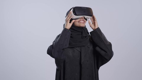 Enjoyment and entertainment while watching through virtual reality glasses. A dark-skinned Saudi Arabian university student wearing an abaya and hijab, wearing 3D glasses, raises her hand upwards with an expression of integration, gestures of stopping something.