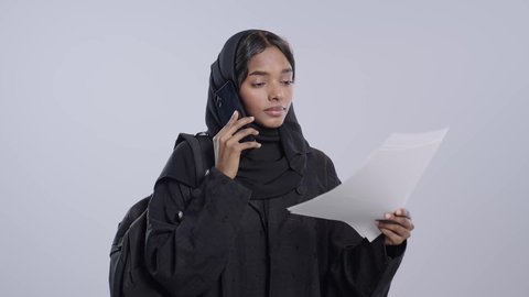اجراء مكالمه هاتفيه ،اعداد التقارير الخاصة ، طالبة جامعية سمراء عربيه سعوديه ترتدي العباءه والحجاب تحمل بيدها مجموعة من الاوراق تمسك بيدها الهاتف المحمول ،  مراجعة ومتابعة الخطط والمشاريع الدراسيه، التدقيق في قراءة الملاحظات، خلفية بيضاء
