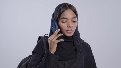 Making a phone call, browsing websites and applications, chatting and talking with friends, a dark-skinned Saudi Arabian university student wearing an abaya and hijab and holding a black bag, using modern technologies in daily life, white background