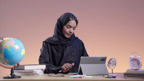استخدام التكنولوجيا الحديثة في الأعمال الدراسيه ،التعامل مع المنصات الاونلاين فى التعليم عن بعد، بورتريه لطالبة جامعية سمراء عربيه سعوديه ترتدي العباءه والحجاب  تجلس على مكتب وتستخدم جهاز التابلت،  دمج التقنيات الحديثة بالمجال التعليمي