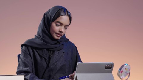 التعامل مع المنصات الاونلاين فى التعليم عن بعد، بورتريه لطالبة جامعية سمراء عربيه سعوديه ترتدي العباءه والحجاب  تجلس على مكتب وتستخدم جهاز التابلت، استخدام التكنولوجيا الحديثة في الأعمال الدراسيه ، دمج التقنيات الحديثة بالمجال التعليمي