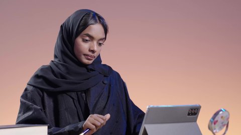 بورتريه لطالبة جامعية سمراء عربيه سعوديه ترتدي العباءه والحجاب  تجلس على مكتب وتستخدم جهاز التابلت، استخدام التكنولوجيا الحديثة في الأعمال الدراسيه ، دمج التقنيات الحديثة بالمجال التعليمي ،التعامل مع المنصات الاونلاين فى التعليم عن بعد